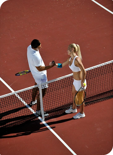 Two tennis players, a man and a woman, are shaking hands over the net on a clay court. Both are dressed in white, conveying a sense of sportsmanship.