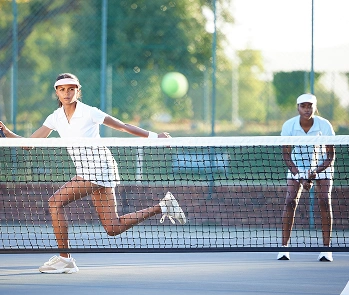 Two women play tennis on an outdoor court. The player in the foreground is mid-swing, focused on the ball, while her partner waits behind her.