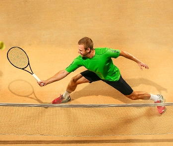 A male tennis player in a green shirt and black shorts lunges at the net on a clay court, stretching to hit a ball. The scene conveys action and intensity.