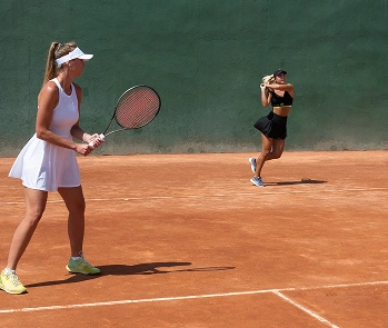 Two women play tennis on a clay court. The woman in white stands ready in the foreground, while the woman in black strikes a backhand in the background.