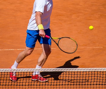 A tennis player in white and blue attire is about to strike a yellow tennis ball with a racquet on a clay court. The scene conveys concentration and action.