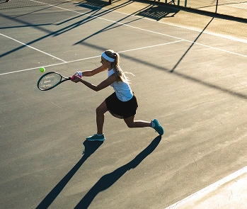 A female tennis player in action, wearing a headband, white top, and black skirt. She is focused on hitting a ball on a sunlit court.