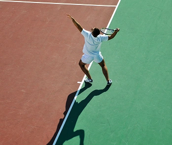 Overhead view of a tennis player in white serving on a green and red court. The player's shadow stretches across the court, capturing a dynamic motion.